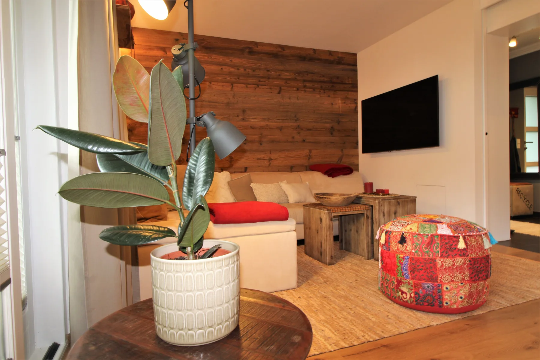 A cozy living room with seating, a wall-mounted television, and a colorful round ottoman.