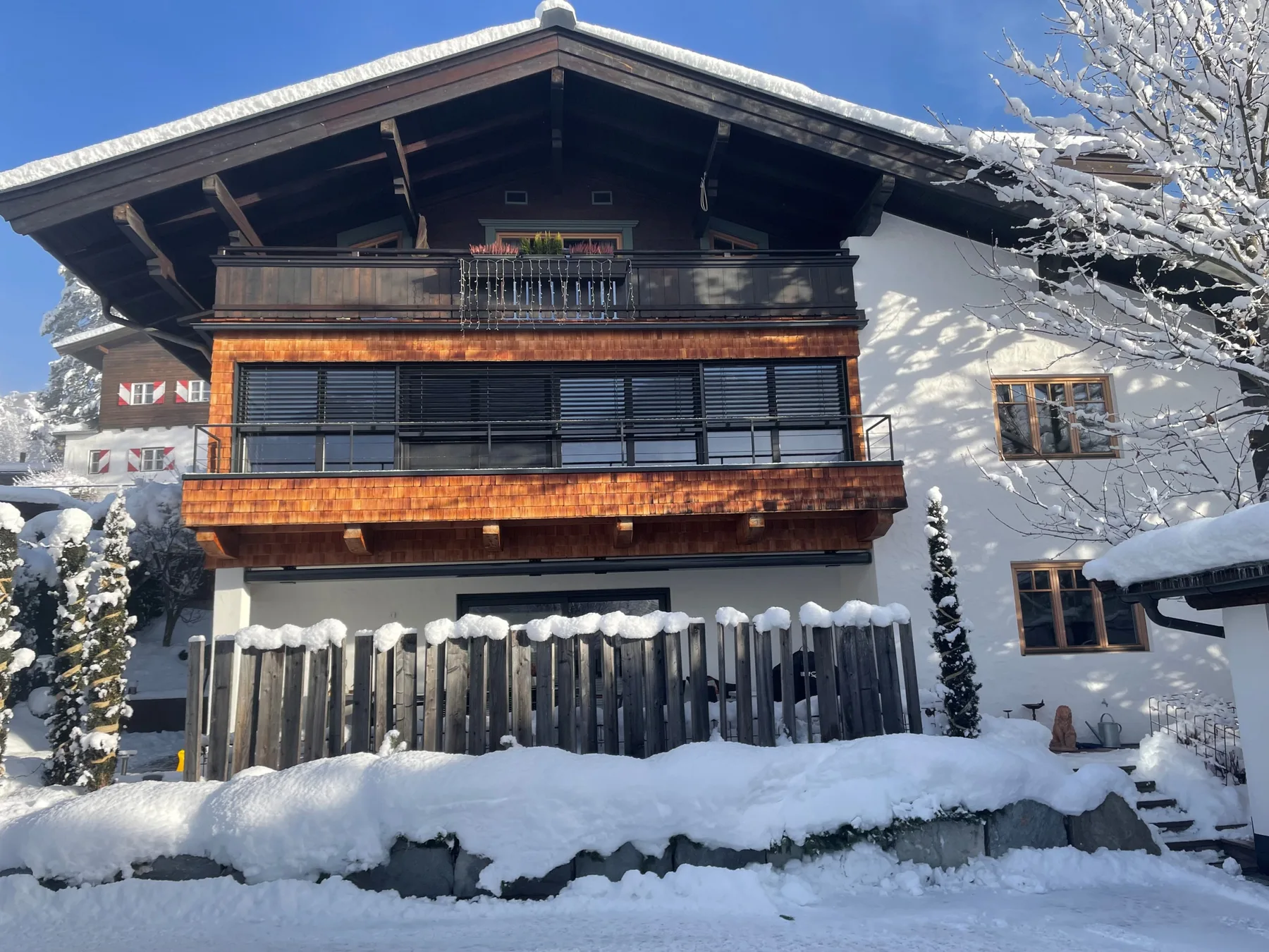 A snow-covered alpine house with wooden balconies stands beneath a clear blue sky.