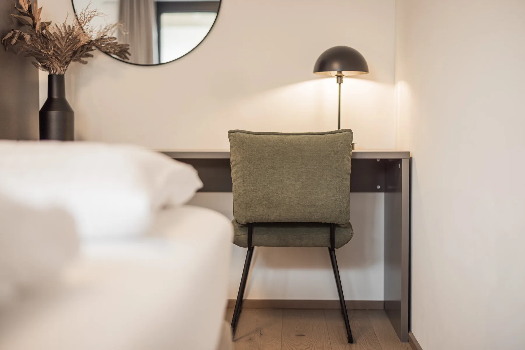 A bedroom workspace features a desk, chair, and lamp beside the bed.