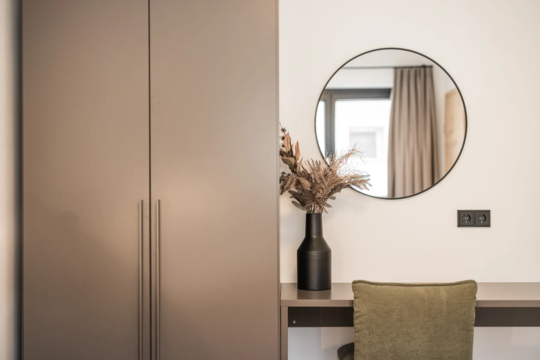 A bedroom dressing area with a wardrobe, desk, and round wall mirror.