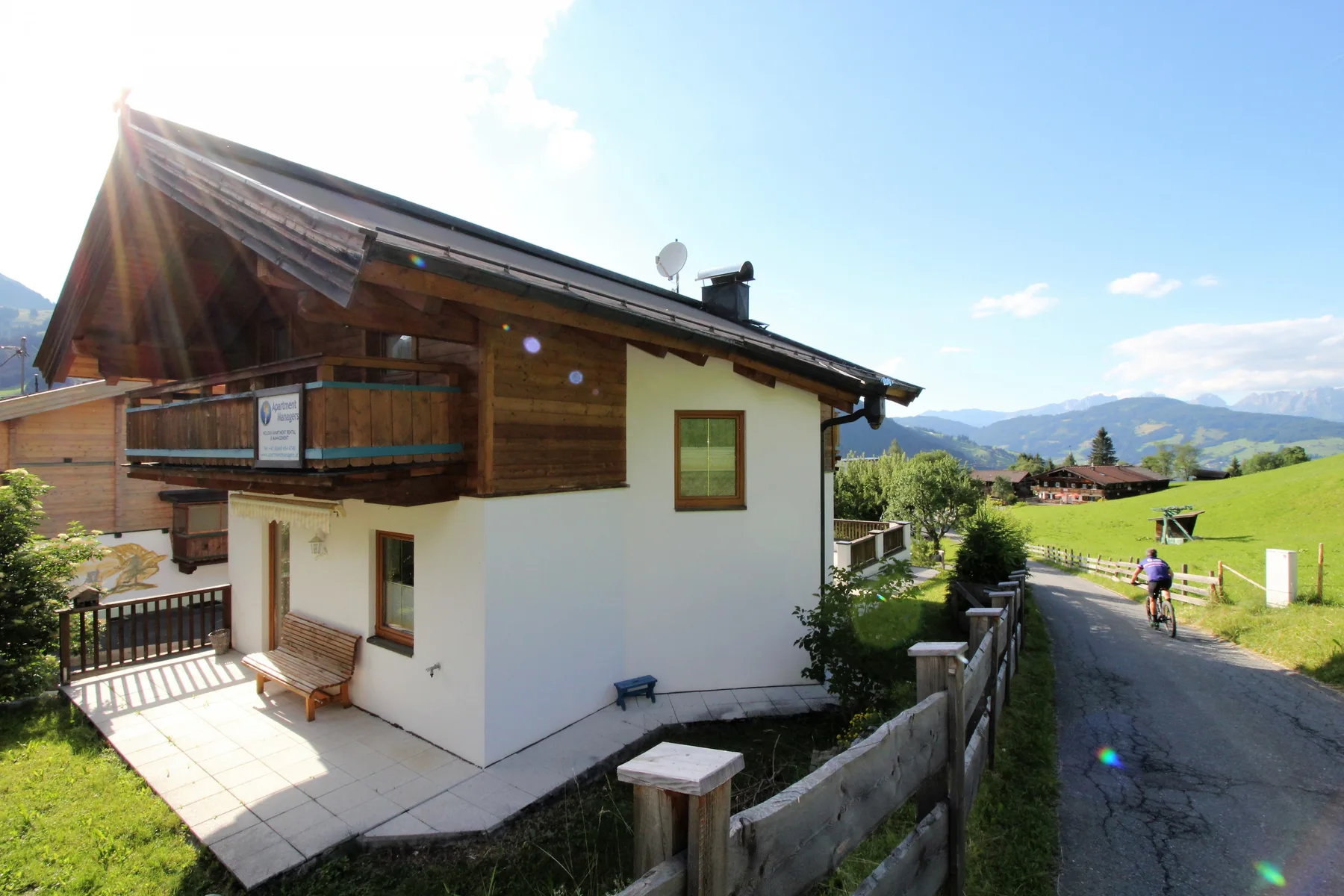 A chalet-style house beside a paved lane, with green hills and mountains beyond.
