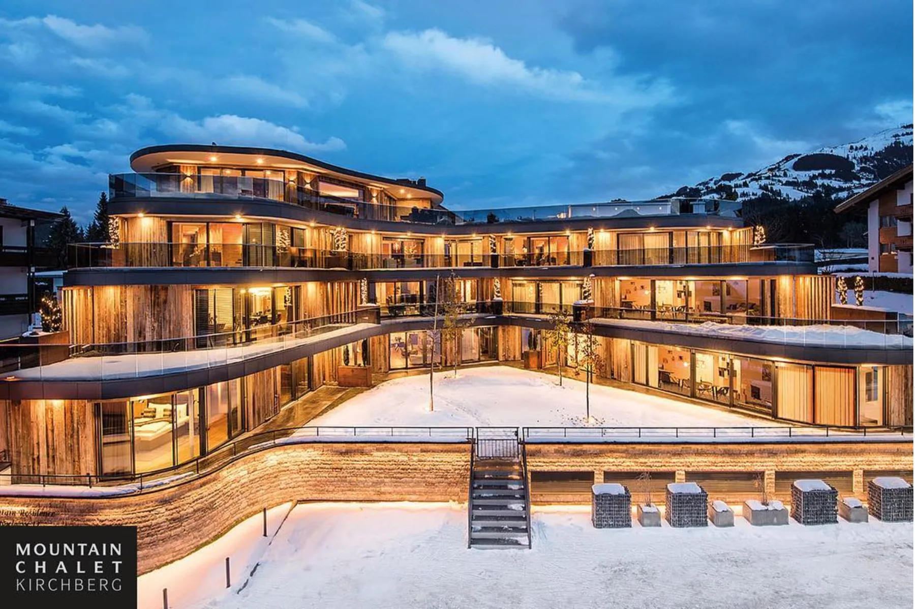 Un complejo de chalets modernos se ilumina al anochecer junto a un patio cubierto de nieve.
