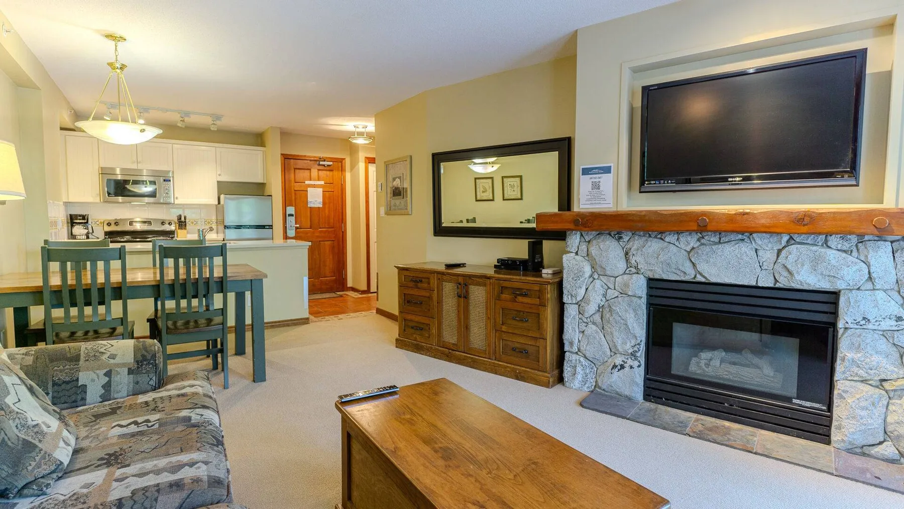 A living room with a stone fireplace, television, and adjoining kitchen.