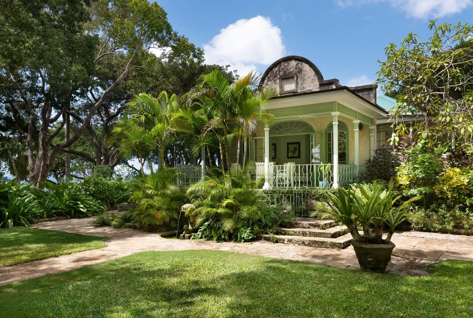 A pale green house with a columned porch sits amid lush garden greenery.