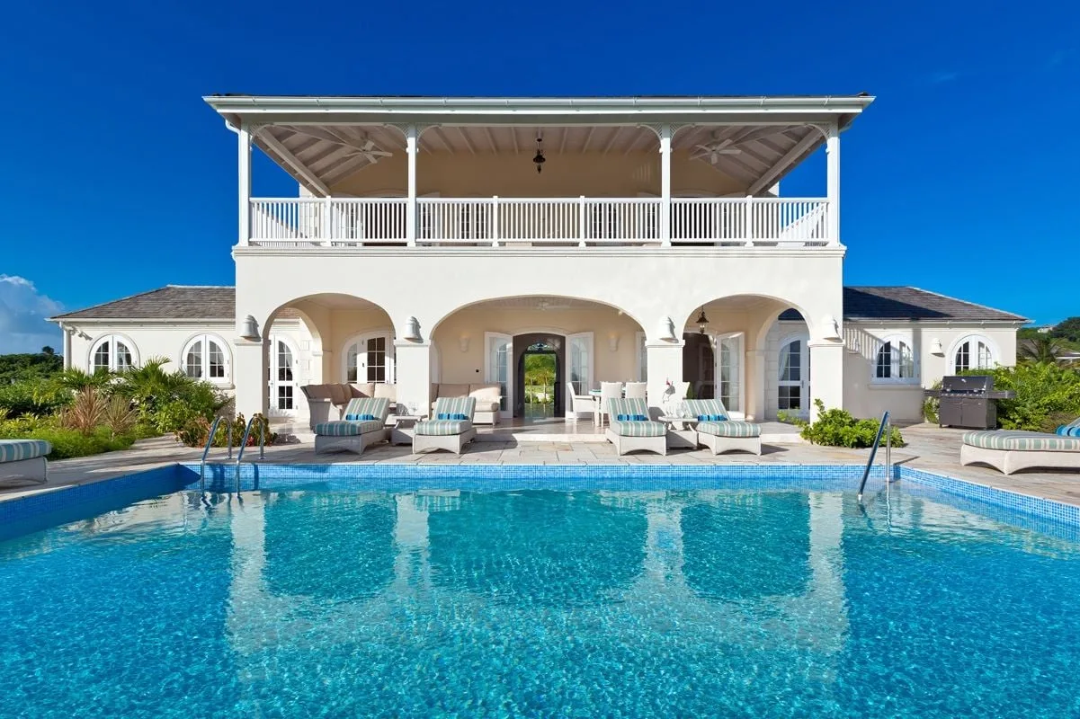 Una gran villa blanca da a una piscina, con asientos cubiertos en el patio.