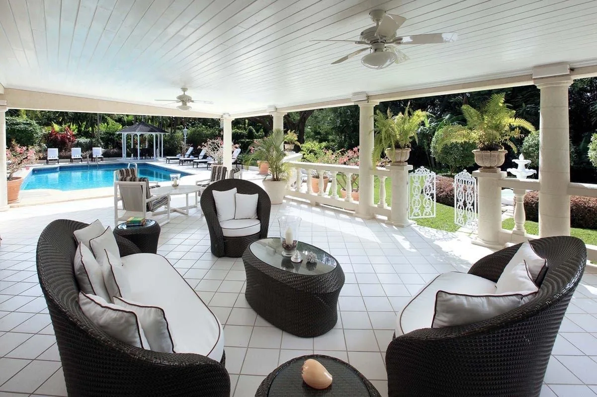 Un patio cubierto con asientos acolchados da a una piscina y un jardín.