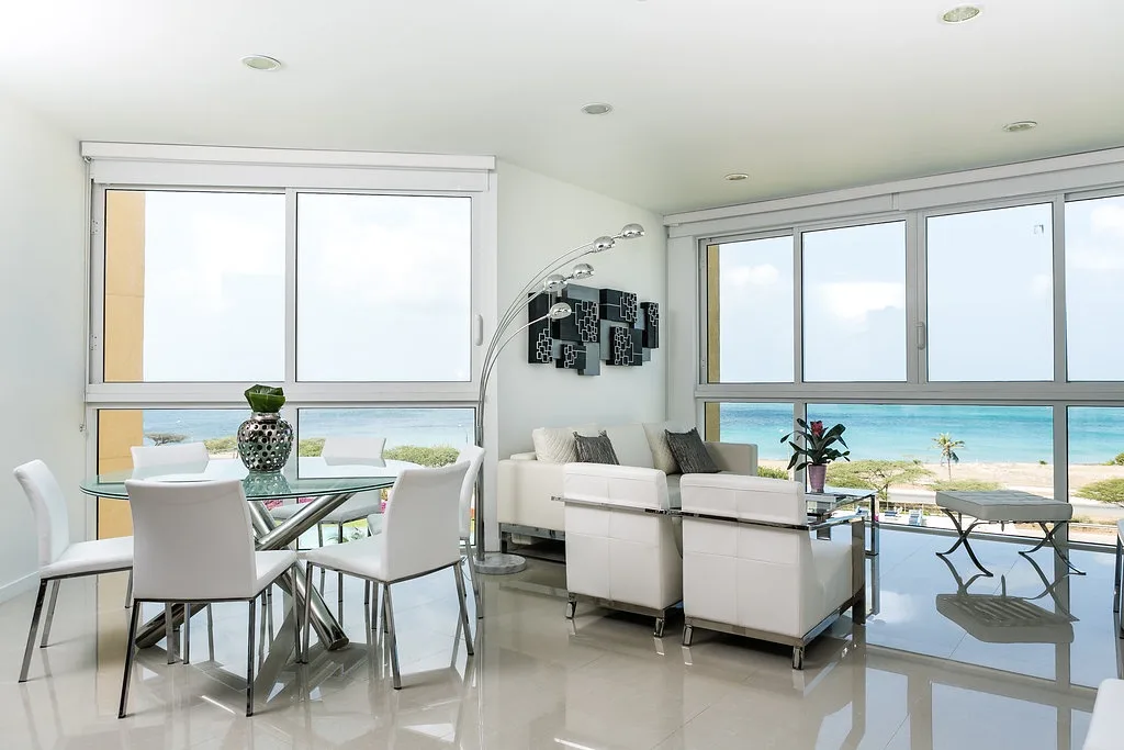La luz del sol llena tu moderna sala de estar y comedor, donde los ventanales del piso al techo enmarcan impresionantes vistas de la playa y las suaves olas más allá.