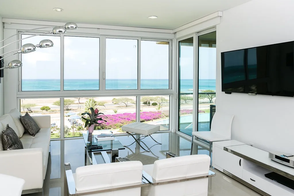 Relájate en tu luminosa sala de estar con impresionantes vistas al mar. Los ventanales del piso al techo enmarcan jardines coloridos y aguas turquesas resplandecientes, mientras que el mobiliario moderno crea un espacio sereno y cómodo para tu estancia junto a la costa.