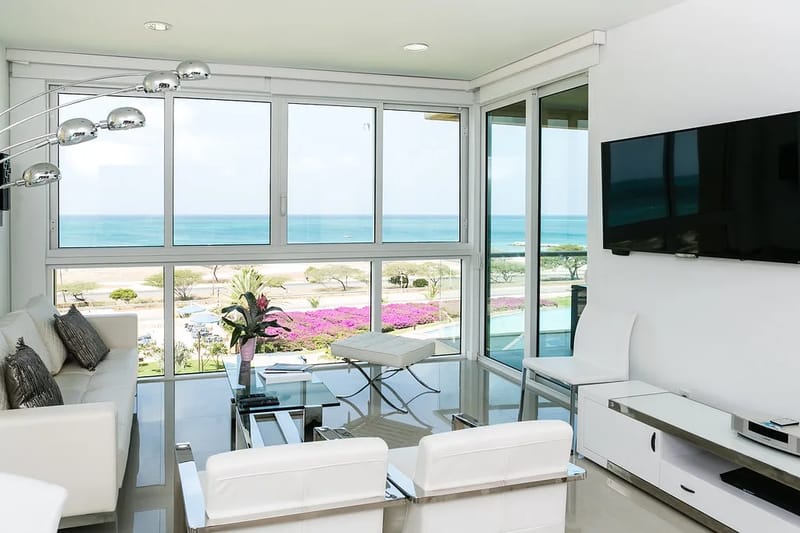 Relájate en tu luminosa sala de estar con impresionantes vistas al mar. Los ventanales del piso al techo enmarcan jardines coloridos y aguas turquesas resplandecientes, mientras que el mobiliario moderno crea un espacio sereno y cómodo para tu estancia junto a la costa.