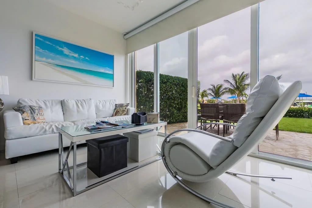 Sink into the plush white sofa or sleek rocking chair as natural light floods your space through floor-to-ceiling windows framing lush tropical views and the nearby pool.