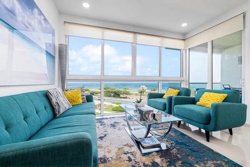 Relájate en esta luminosa sala de estar con cómodos asientos acolchados de color verde azulado, ventanas de piso a techo que enmarcan vistas al mar y un ambiente acogedor perfecto para descansar después de tus aventuras en la playa.