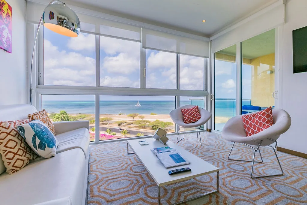 Imagínate descansando en esta luminosa sala de estar, donde los ventanales de piso a techo enmarcan impresionantes vistas al mar. Los elegantes muebles modernos y los toques de color coral crean un ambiente costero relajado, perfecto para tu escapada junto a la playa.