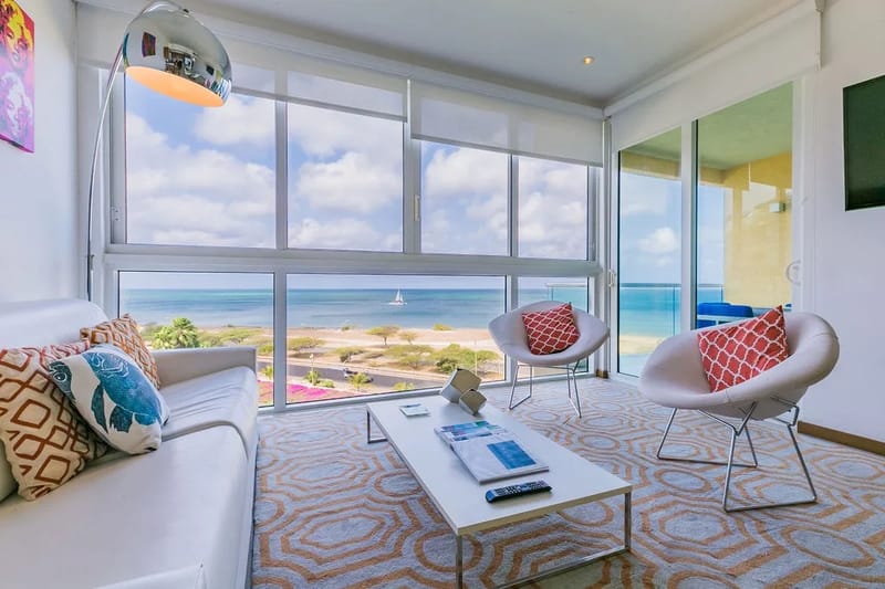 Imagínate descansando en esta luminosa sala de estar, donde los ventanales de piso a techo enmarcan impresionantes vistas al mar. Los elegantes muebles modernos y los toques de color coral crean un ambiente costero relajado, perfecto para tu escapada junto a la playa.