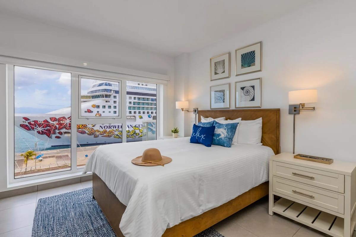 A coastal bedroom where crisp white linens and nautical accents frame stunning harbor views through floor-to-ceiling windows, inviting peaceful mornings with the water just beyond your balcony.
