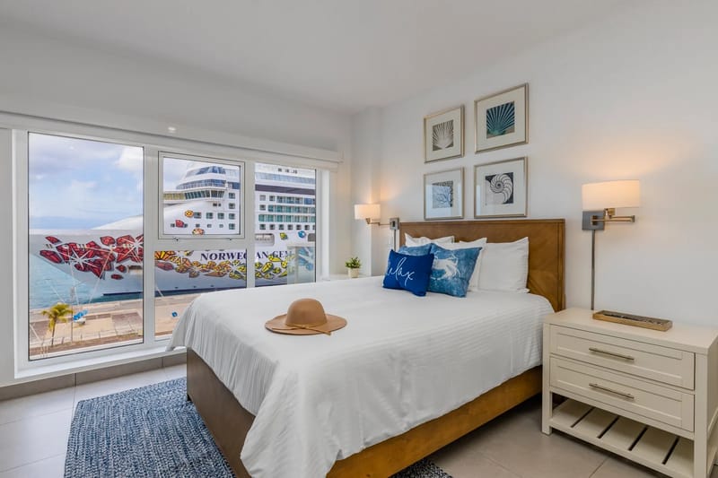 A coastal bedroom where crisp white linens and nautical accents frame stunning harbor views through floor-to-ceiling windows, inviting peaceful mornings with the water just beyond your balcony.