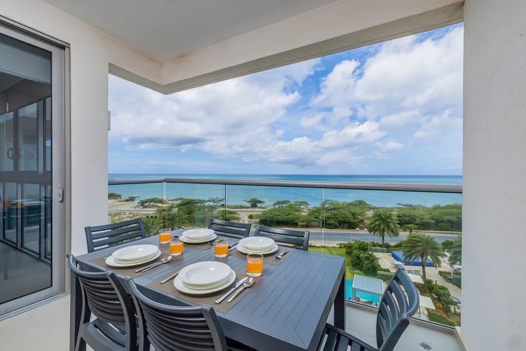 Coastal property balcony with panoramic ocean views, poolside setting, and tropical landscape surroundings. Open-air dining space overlooks turquoise waters and lush vegetation.