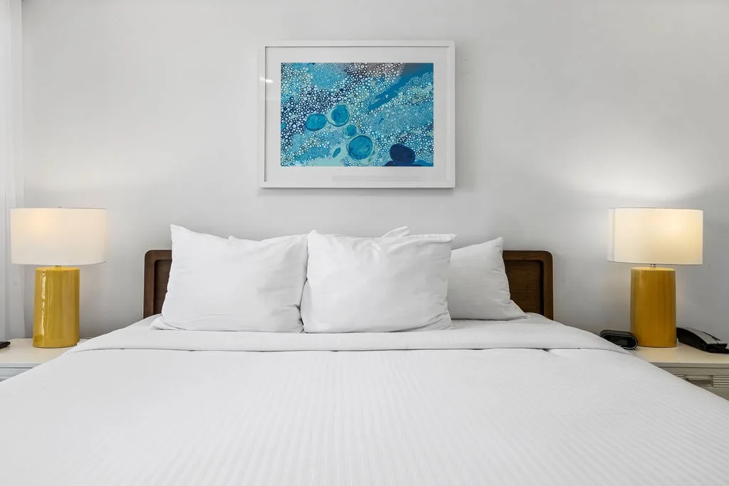 Dormitorio con ropa de cama blanca impecable, arte inspirado en el océano e iluminación cálida junto a la cama.