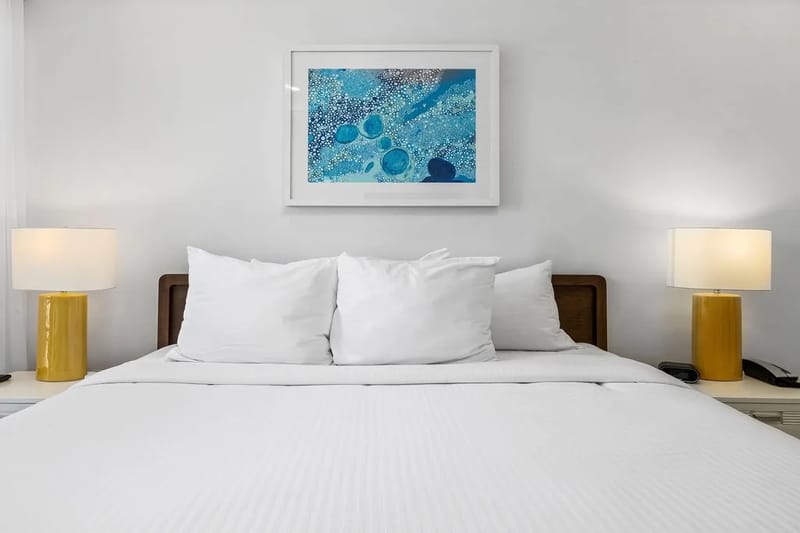Dormitorio con ropa de cama blanca impecable, arte inspirado en el océano e iluminación cálida junto a la cama.
