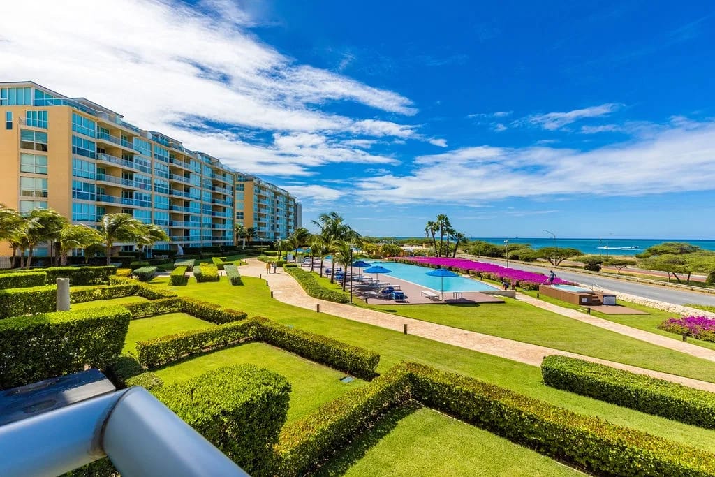 Impresionante complejo vacacional frente a la playa con jardines bien cuidados, una piscina resplandeciente y amplias vistas al mar, enmarcado por un vibrante paisaje tropical y cielos azules infinitos.