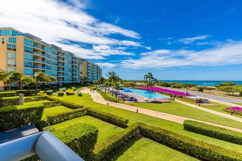 Impresionante complejo vacacional frente a la playa con jardines bien cuidados, una piscina resplandeciente y amplias vistas al mar, enmarcado por un vibrante paisaje tropical y cielos azules infinitos.