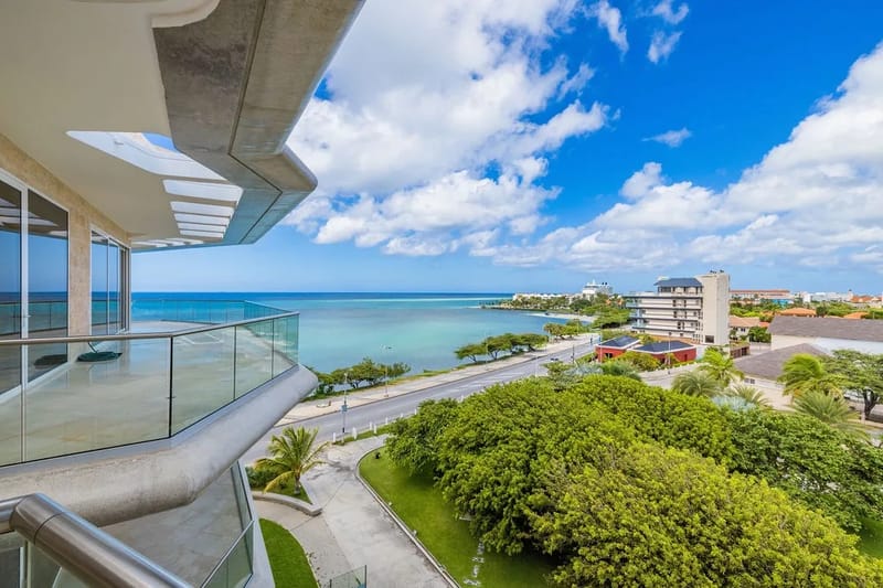 A modern property balcony offers sweeping sea views with lush coastal surroundings, situated near calm turquoise waters and vibrant tropical greenery in a peaceful beachside neighborhood.