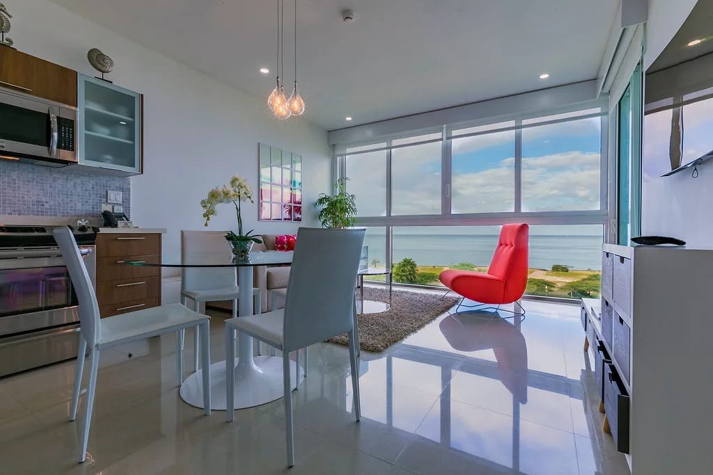 Luz del sol a través de ventanas de piso a techo, una mesa de comedor y vistas al mar.