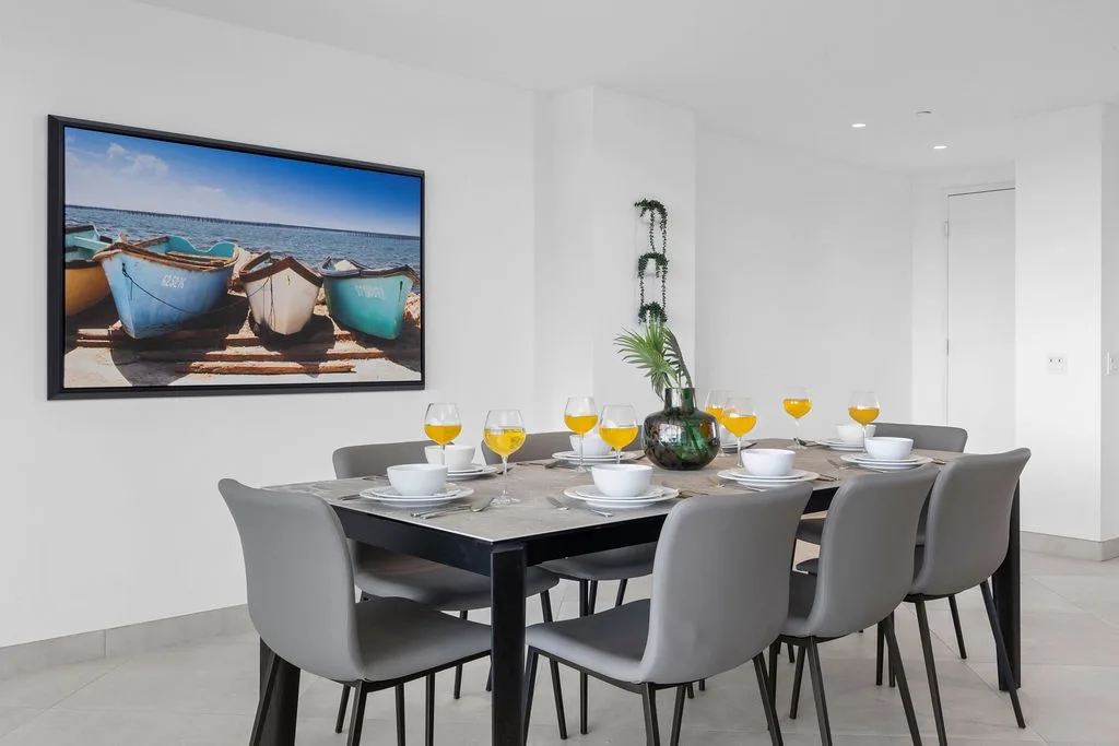 Zona de comedor con una mesa puesta para ocho personas y obras de arte de temática costera.