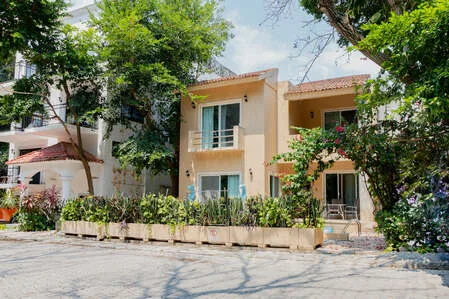 Un edificio residencial de dos plantas se alza detrás de árboles frondosos y plantas de jardín.