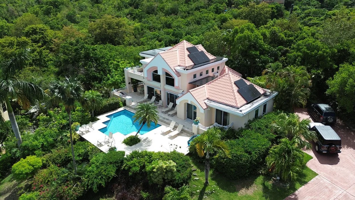 A large house beside a swimming pool, surrounded by dense greenery.
