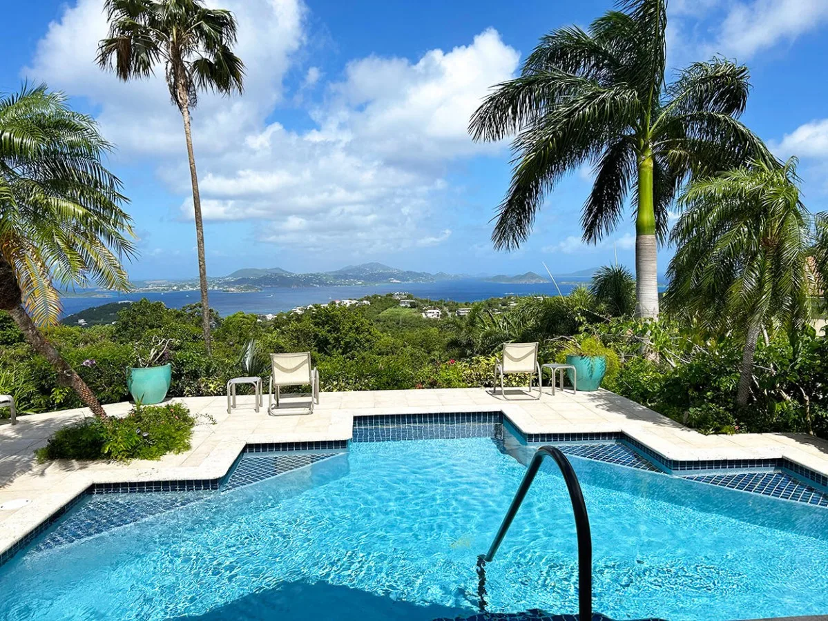 A swimming pool overlooks tropical greenery and distant islands beneath a blue sky.
