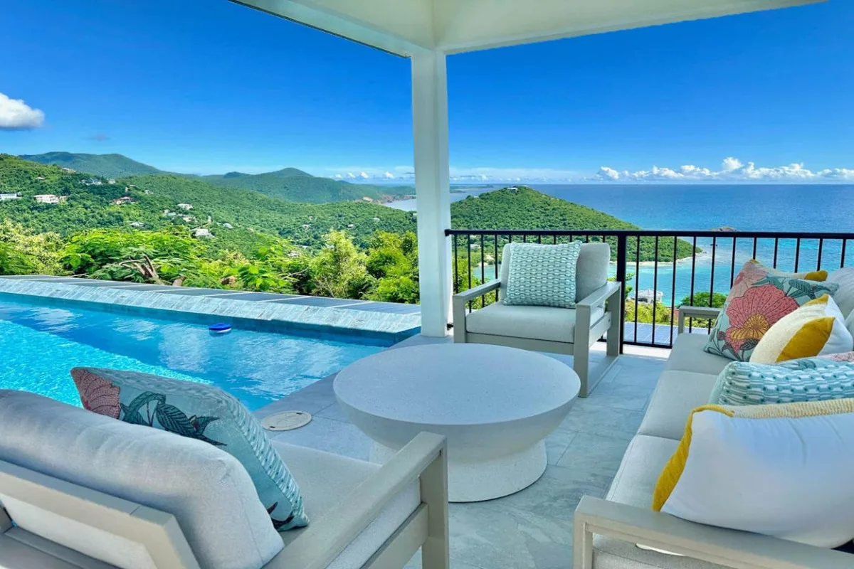An outdoor seating area overlooks a swimming pool, green hills, and the sea.