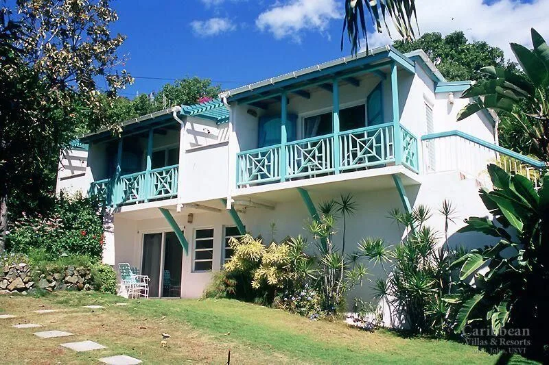 Una casa blanca de dos plantas con balcones turquesa está rodeada de plantas tropicales.