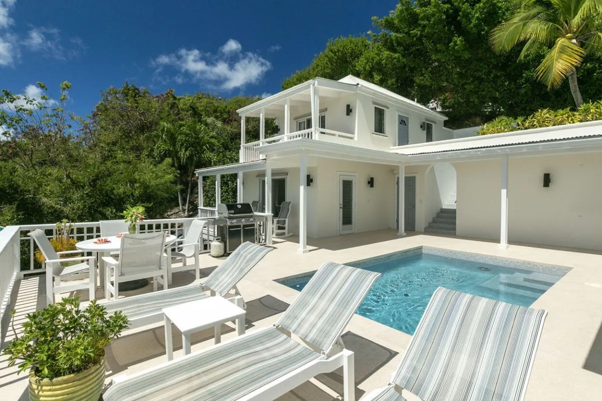 Una casa blanca se abre a una terraza junto a la piscina con asientos para descansar.