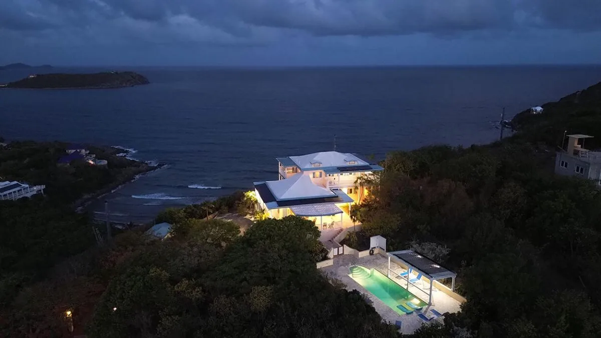 A hillside property overlooks the sea at dusk, with illuminated buildings and a pool.