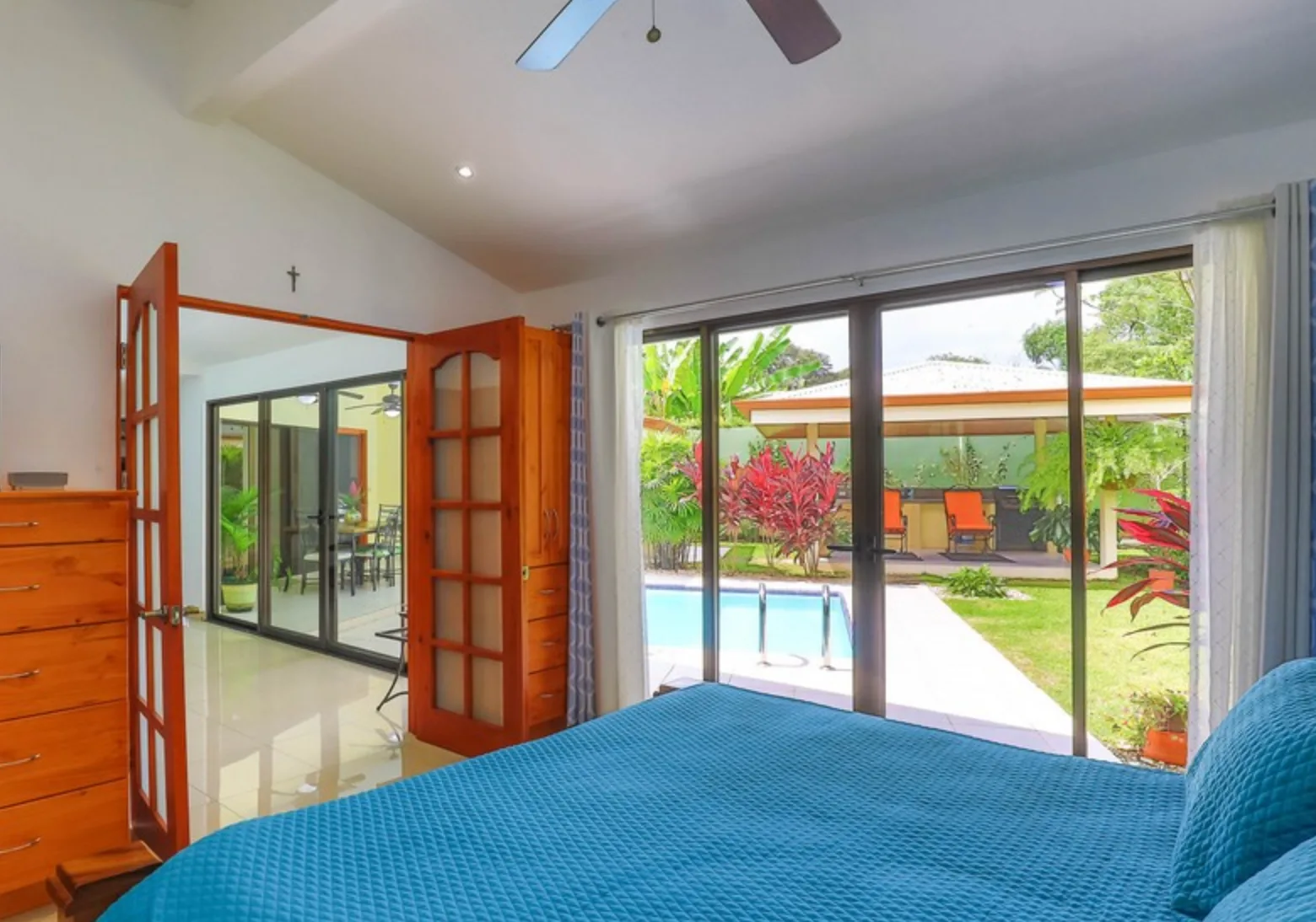 Bedroom with a blue bedspread and sliding glass doors overlooking a pool and garden.