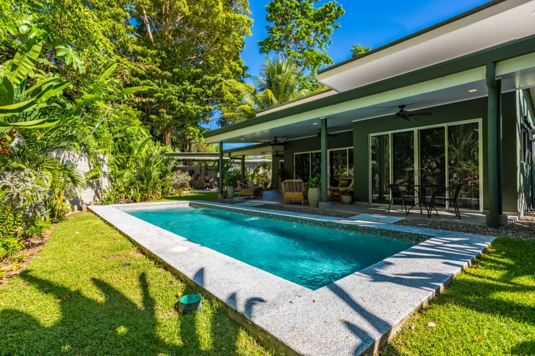 Una piscina junto a una casa moderna, con un patio cubierto y vegetación alrededor.