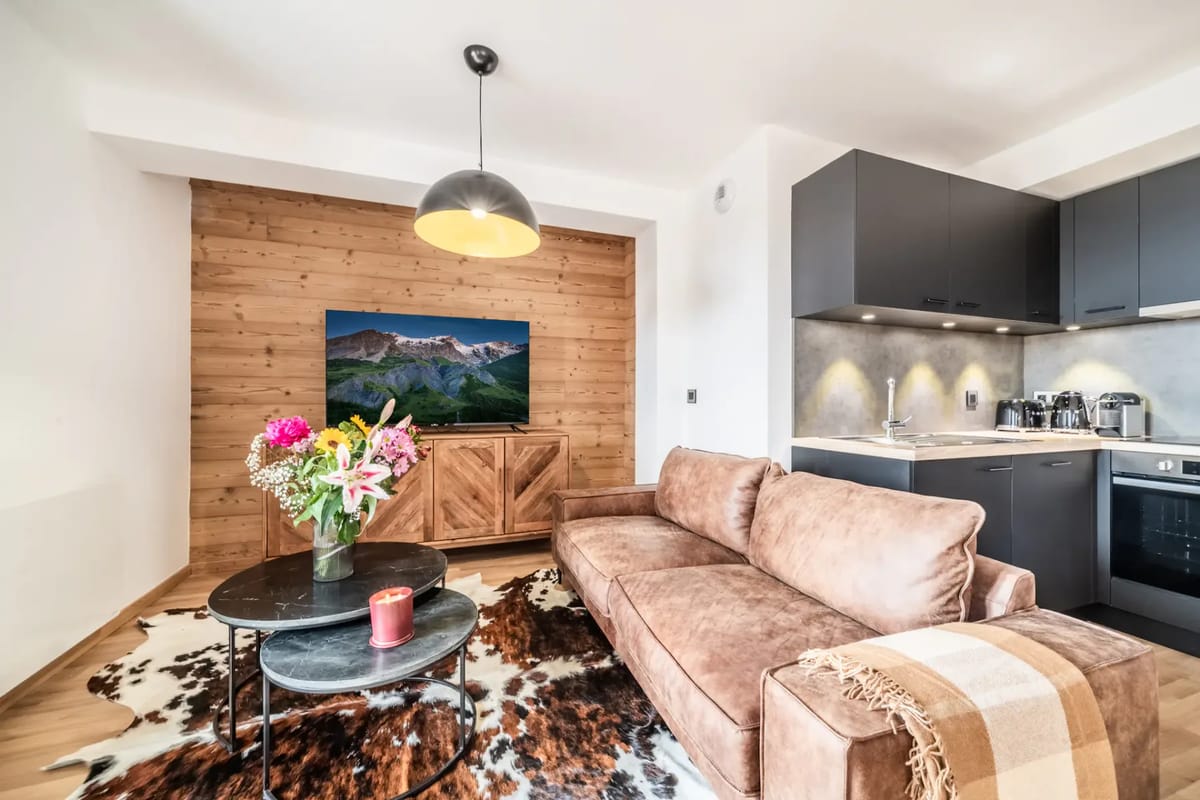 Salón moderno con un sofá marrón, zona de comedor, televisor montado en la pared y cocina compacta.