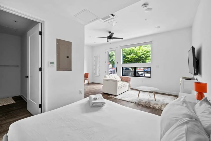Dormitorio y área de estar luminosos con cama, sofá, televisor y ventanas grandes.