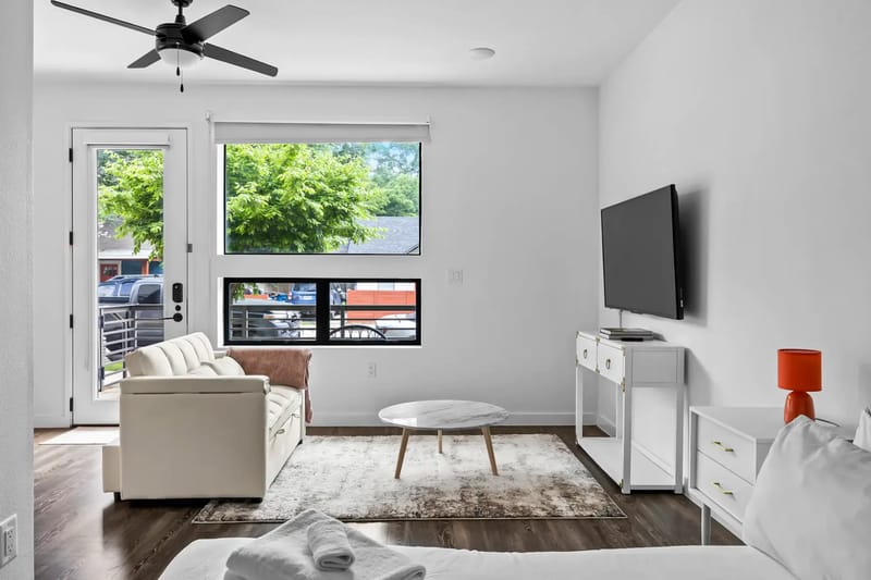 Sala moderna con paredes blancas, sofá, televisor, lámpara naranja, piso de madera y ventilador de techo.