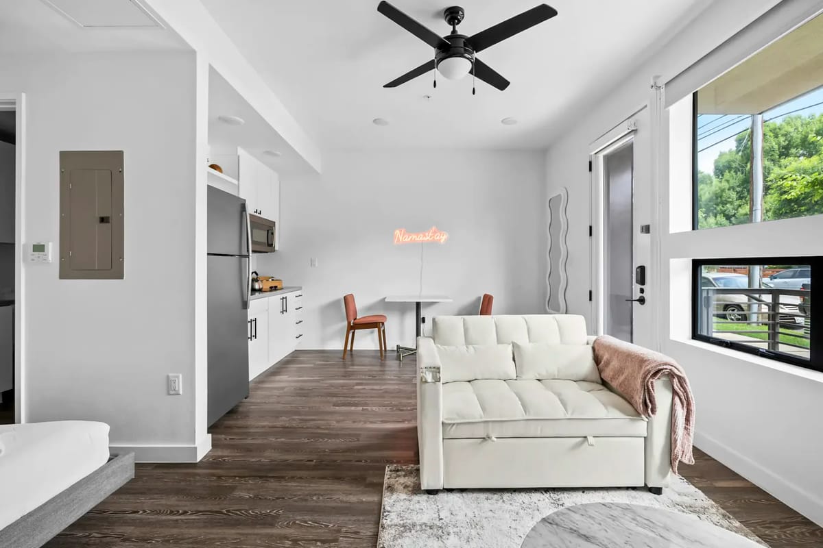Área de estar moderna con sofá blanco, ventilador de techo, ventanas grandes, una pequeña cocina y mesa de comedor.