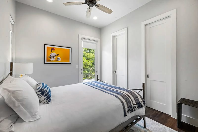 Cozy bedroom with a striped blanket, wall art, ceiling fan, and leafy outdoor view.