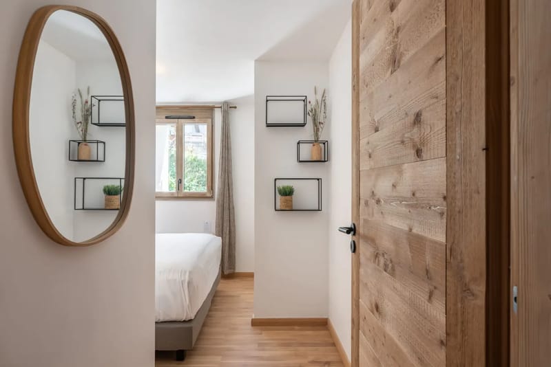 Un dormitorio minimalista con una puerta de madera, un espejo redondo y pequeñas plantas en macetas sobre estantes de pared.