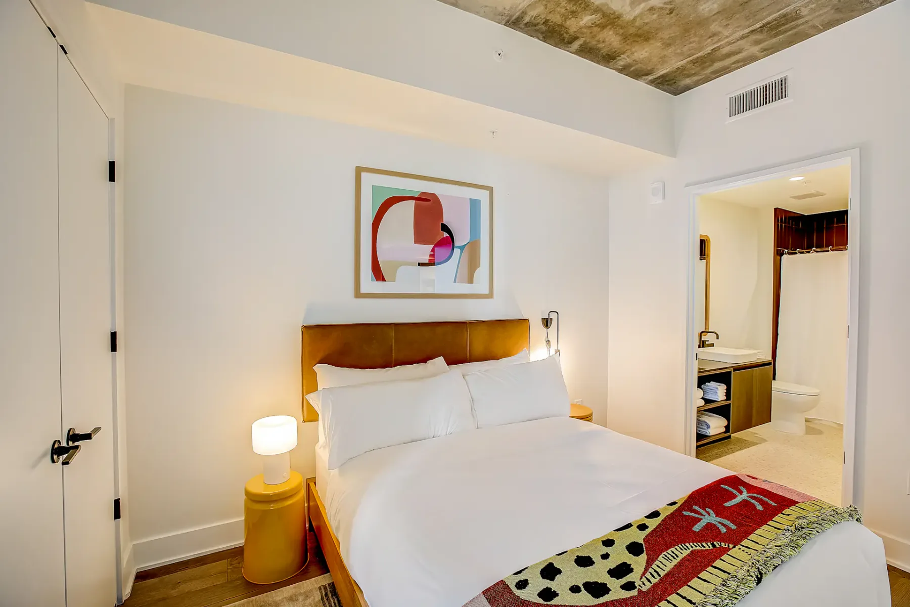 Dormitorio con cama doble, ropa de cama blanca, obras de arte abstracto y acceso a un baño.
