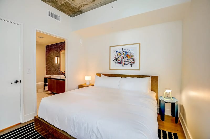 Dormitorio con cama grande, ropa de cama blanca, obras de arte y una puerta a un baño contiguo.