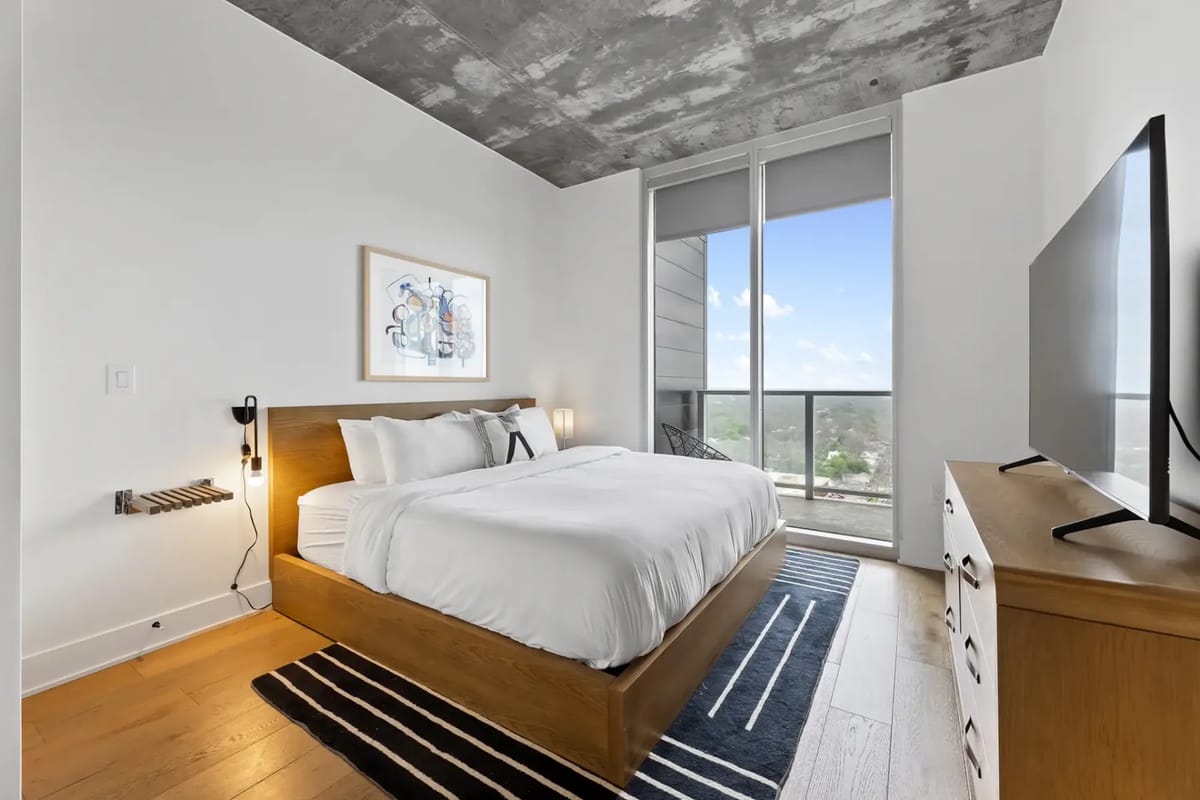 Modern bedroom with a large bed, wood furniture, wall art, and a tall window.