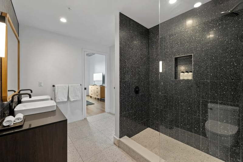 Baño moderno con una ducha revestida de azulejos oscuros, dos lavabos y toallas.