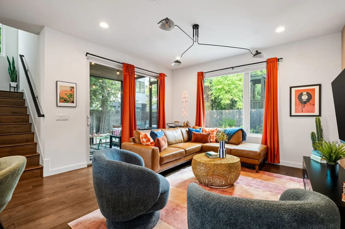 Sala de estar moderna con sofá de cuero marrón, sillas giratorias azules, cojines naranjas y azules y un área de comedor. Grandes ventanas se abren hacia un patio.