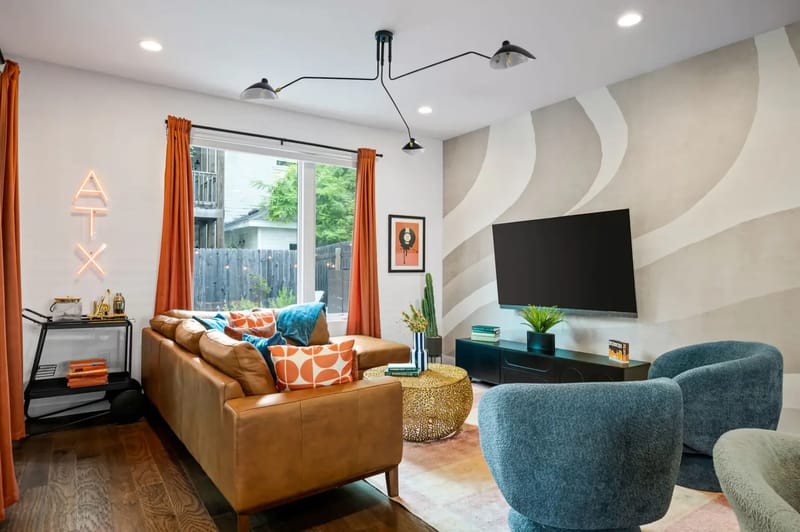 Modern living room with orange and blue seating, vinyl record decor, TV, orange curtains, and a neon