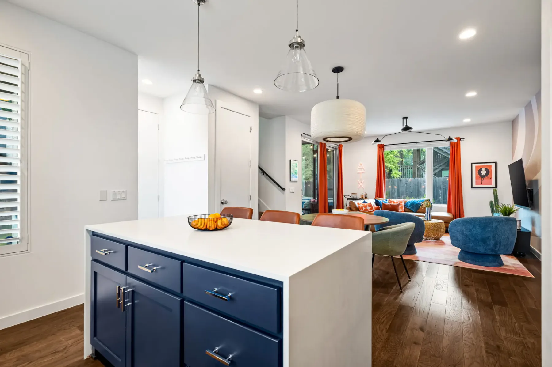 Área de estar moderna de concepto abierto con una isla de cocina, gabinetes azul marino, asientos coloridos, pisos de madera y detalles en naranja brillante.