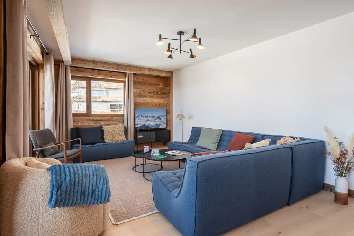 Modern living room with a blue sectional sofa, armchairs, wooden accents, and a coffee table on a beige rug. Large windows provide ample light.